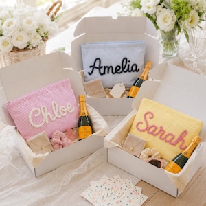 Può includere: Tre scatole regalo bianche contenenti pochette personalizzate con i nomi Chloe, Amelia e Sarah in corsivo. Ogni scatola include una piccola bottiglia di champagne, un portagioie e un elastico per capelli. Le pochette sono a righe rosa, blu e gialle.