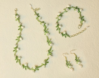 May include: A jewelry set featuring a necklace, bracelet, and earrings. The set is designed with green enamel leaves, white flower accents, and faux pearls. The necklace and bracelet have a gold-tone chain. The earrings have a gold-tone hook.