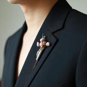 Puede incluir: Un prendedor plateado con un pequeño ramo de flores rosas secas y vegetación. El prendedor está sujeto a un blazer azul marino oscuro, creando un accesorio elegante y único. El arreglo floral añade un toque de naturaleza al atuendo formal.