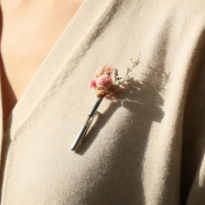 Puede incluir: Un broche plateado con un pequeño ramo de flores secas rosas. Las flores están sujetas en un soporte metálico rectangular y delgado. El broche está prendido a un suéter beige, creando un accesorio delicado y natural.