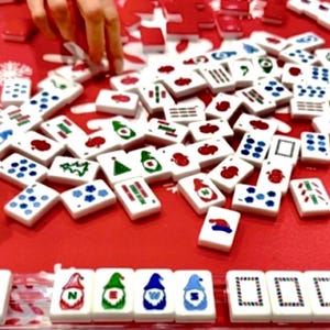 May include: A red surface is covered with white Mahjong tiles, some with Christmas-themed designs. Hands are visible, arranging the tiles. The tiles feature various symbols and colors, including red, green, and blue. The game is set up for play.