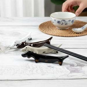May include: Four ceramic dachshund-shaped chopstick rests in white, brown, and black. A pair of black chopsticks rests on the table. A white bowl with blue floral designs and a matching spoon sit on a woven mat.