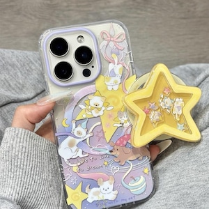 May include: Clear phone case with a whimsical design featuring cartoon animals, stars, and a rainbow. The case has a purple border and a yellow star-shaped phone grip. Text on the case reads "to love to dream".