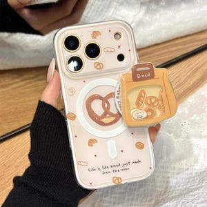 Może przedstawiać: Przezroczyste etui na telefon z motywem pieczywa. Etui posiada ilustracje precli, rogalików i chleba, z napisem "Life is like bread just made from the oven". Do etui dołączony jest uchwyt pop-socket w kształcie chleba.