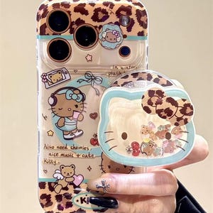Puede incluir: Funda transparente para tel&eacute;fono con dise&ntilde;o de estampado de leopardo y tem&aacute;tica de Hello Kitty. La funda presenta un agarre emergente con un dise&ntilde;o de Hello Kitty y una secci&oacute;n transparente con dijes flotantes. El texto en la funda dice "Also need cherries, nice music + cute Kitty."
