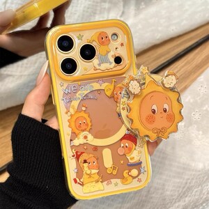 May include: Yellow phone case with a cartoon sun and character design. The case has a clear back with a yellow border and a pop-up sun-shaped phone grip. The case features the text "We are Twinkle Twinkle".