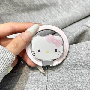 May include: A white and pink phone ring holder featuring the Hello Kitty character. The ring holder has a pink polka dot border and a silver hinge. The text "Y2k x Cases" is visible in the background.