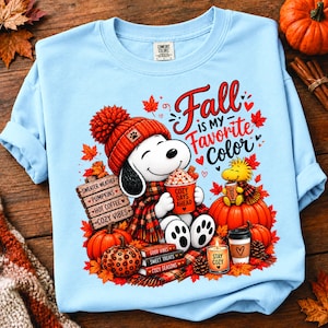 Puede incluir: Una sudadera azul claro de cuello redondo con un gráfico de Snoopy usando un gorro y bufanda de punto naranja, sosteniendo una taza con la etiqueta "COZY DAYS AHEAD". El diseño incluye calabazas, libros, una vela y el texto "Fall IS MY FAVORITE Color."