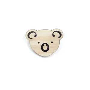 Könnte beinhalten: Ein ausgeschnittener Koala-Kopf aus Holz mit schwarzen, bemalten Merkmalen. Der Koala hat große Ohren, eine kleine Nase und einen breiten Mund.