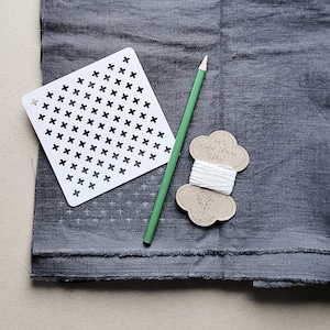 May include: A still-life image featuring a white cross-stitch stencil, a green pencil, and a spool of white thread labeled "100% Linen Sashiko Yarn." These items rest on a textured, dark gray fabric, suggesting a crafting or sewing project.