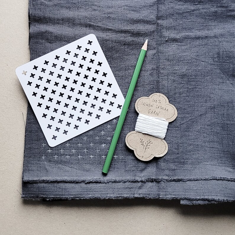 May include: A still-life image featuring a white cross-stitch stencil, a green pencil, and a spool of white thread labeled "100% Linen Sashiko Yarn." These items rest on a textured, dark gray fabric, suggesting a crafting or sewing project.