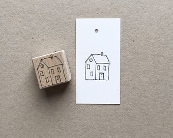 Puede incluir: Un sello de goma de madera con un dibujo de línea negra de una casa con dos ventanas y una puerta. El sello está sobre una superficie marrón junto a una tarjeta blanca con la misma imagen.