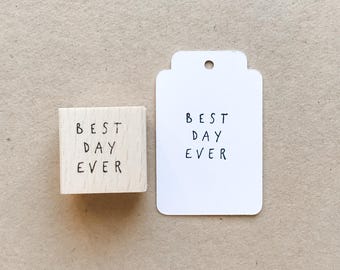 Puede incluir: Un sello de madera y una etiqueta blanca con la frase escrita a mano en negro "BEST DAY EVER". El sello es un pequeño bloque cuadrado. La etiqueta es rectangular con una parte superior redondeada y un agujero.