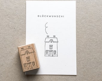 Puede incluir: Un sello de madera con una casa junto a una tarjeta con una ilustración de una casa y el texto "GLÜCKWUNSCH!". El sello y la tarjeta presentan un sencillo dibujo lineal negro de una casa con chimenea y humo. La tarjeta es blanca.