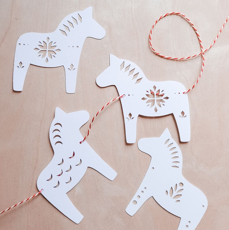 May include: Four white paper horse cutouts with decorative snowflake and floral designs are strung on a red and white twine. The horses are arranged on a light brown wooden surface, creating a festive and handcrafted aesthetic.