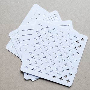 May include: A stack of white stencils with various cut-out patterns. The designs include dots, lines, and cross-shaped floral motifs. The stencils are square with rounded corners, and are stacked on a neutral surface.