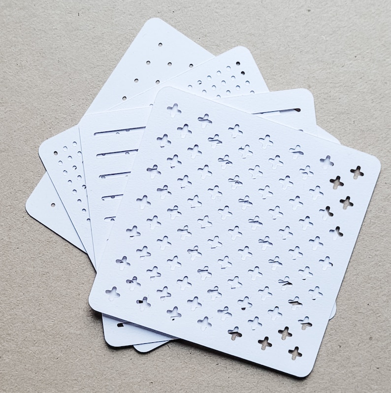 May include: A stack of white stencils with various cut-out patterns. The designs include dots, lines, and cross-shaped floral motifs. The stencils are square with rounded corners, and are stacked on a neutral surface.