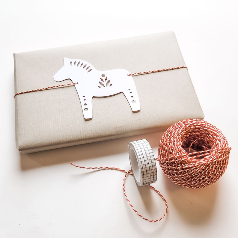 May include: A wrapped gift with a white horse-shaped tag, tied with red and white twine. A roll of grid-patterned tape and a ball of twine are next to the gift. The gift is wrapped in light brown paper.