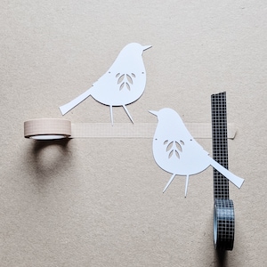 以下が含まれることがあります： 白い紙の鳥の切り抜き2つ。葉のディテールがあり、ベージュの表面に配置され、ベージュのグリッドテープで接続されています。ベージュのグリッドテープと黒のグリッドテープのロールも存在します。この画像は、ミニマリストで工芸品のような美学を持っています。