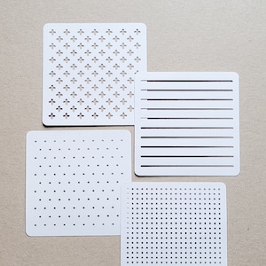 May include: Four white stencils with different patterns. One stencil has a repeating cross design, another has horizontal lines, and two have dot patterns of varying densities. The stencils are square with rounded corners.
