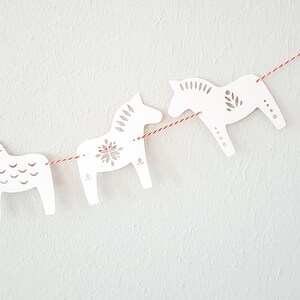 May include: A garland of white wooden Dala horses strung on a red and white twine. The horses have decorative cut-out patterns and are arranged against a white textured background. The garland is a festive decoration.