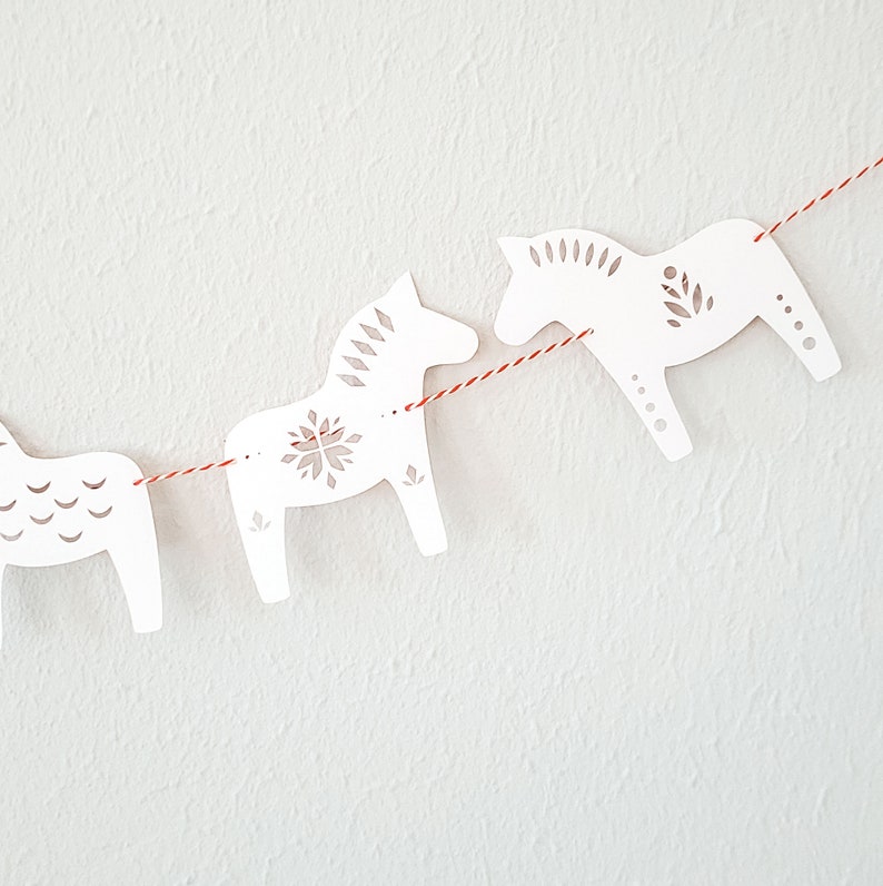 May include: A garland of white wooden Dala horses strung on a red and white twine. The horses have decorative cut-out patterns and are arranged against a white textured background. The garland is a festive decoration.