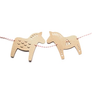 May include: Two tan-colored, cutout wooden horse ornaments are strung on a red and white twine. The horses have decorative cutouts and are set against a white background. The horses are facing each other.