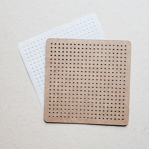 May include: Two square pegboards, one white and one brown, are displayed. Both pegboards have a grid of evenly spaced holes. The white pegboard is slightly larger and positioned behind the brown pegboard. The pegboards are likely used for crafts or creating pixel art.