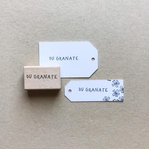 May include: A wooden stamp and two white tags, all bearing the text "DU GRANATE" in black. One tag features a floral pattern. The items are arranged on a light brown surface.