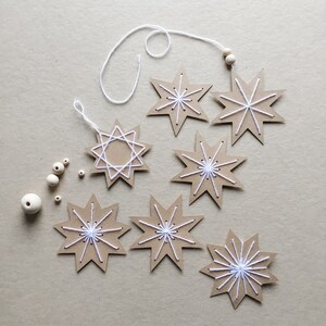May include: Handmade star-shaped ornaments crafted from brown cardboard, adorned with white thread stitching. The stars are connected by white string and wooden beads, perfect for holiday decor. Several loose wooden beads are scattered nearby.