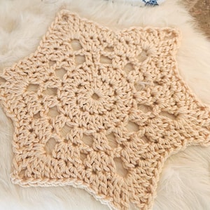 Può includere: Un centrino all'uncinetto a forma di stella di colore crema chiaro. Il pezzo presenta un intricato motivo traforato con un design circolare centrale che si irradia verso l'esterno in cinque punte distinte, mostrando una trama in filato.