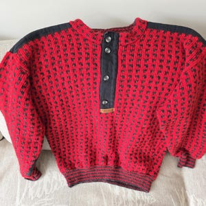 May include: A red and black patterned sweater with a button-up collar. The sweater has black accents on the shoulders and a ribbed hem and cuffs. The sweater is made of a thick, textured knit.