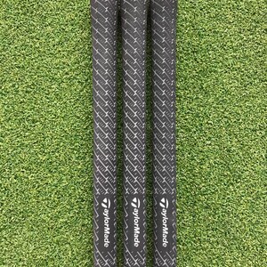 May include: Three golf club grips with black and white patterned grips and red accents. The grips are labeled "Golf Pride" and "TaylorMade". The grips are set against a green background.