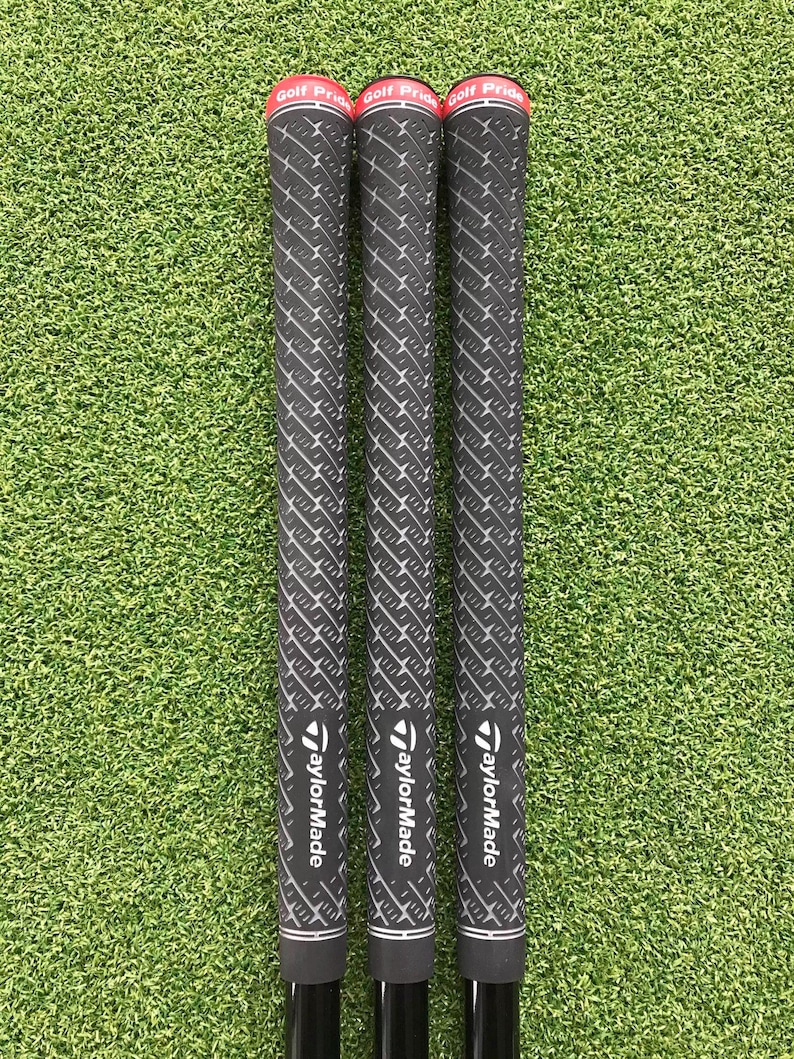 May include: Three golf club grips with black and white patterned grips and red accents. The grips are labeled "Golf Pride" and "TaylorMade". The grips are set against a green background.