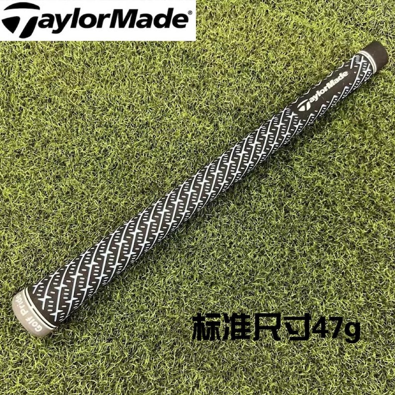 May include: A black and white golf club grip with a textured pattern. The grip has the TaylorMade logo and the text "Golf Pride". The grip is lying on a green grassy surface.