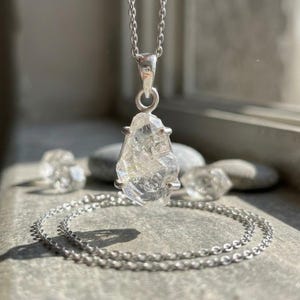 Peut inclure: Un collier en argent avec un pendentif en cristal brut, transparent et à facettes, maintenu par un sertissage à quatre griffes. Le pendentif est suspendu à une fine chaîne en argent, avec d'autres petites pierres de cristal transparentes posées à proximité.