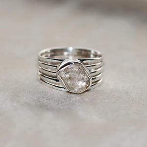 Peut inclure: Bague en argent avec une grosse pierre transparente de forme irrégulière. La bague présente un design à plusieurs bandes, avec cinq bandes parallèles formant la base. La pierre est sertie sur le dessus, créant un bijou unique.
