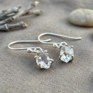 Op de afbeelding: Een paar zilveren oorbellen met ruwe, heldere, gefacetteerde kristallen stenen die op hun plaats worden gehouden door kleine metalen pootjes. De oorbellen hebben gebogen zilveren oorhaken om ze op te hangen en tonen de natuurlijke, grillige vorm.