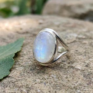 Op de afbeelding: Een zilveren ring met een grote, ovaalvormige maansteen cabochon met een doorschijnend, melkachtig uiterlijk en een subtiele blauwe iriserende glans. De ringband is gemaakt van gepolijst zilver en splitst zich in twee armen bij de zetting.