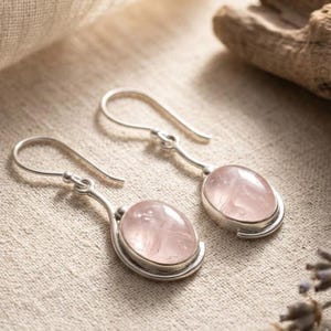 Könnte beinhalten: Ein Paar Ohrhänger mit ovalen, durchscheinenden, blassrosa Rosenquarz-Edelsteinen in silberfarbenen Metallfassungen mit geschwungenen Ohrhaken, die oben an den Steinhalterungen befestigt sind.