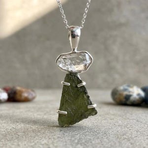 May include: A pendant necklace featuring a clear, faceted crystal stone set above a triangular, textured olive-green moldavite stone. Both stones are held in a silver setting, suspended from a delicate silver chain against a neutral background.