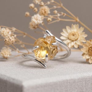 May include: A silver ring featuring a raw, translucent yellow citrine gemstone held in a four-prong setting. The band has a bypass design, with two ends that overlap slightly, creating a sleek and modern look that highlights the natural texture.