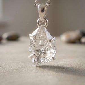 Op de afbeelding: Een heldere, ruwe, veelzijdige kwartskristallen hanger in een gepolijste zilveren zetting met pootjes. De hanger hangt aan een zilveren oogje en is bevestigd aan een fijne zilveren ketting, wat de natuurlijke, onregelmatige textuur van de steen toont.