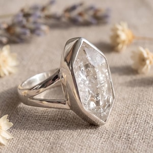 Peut inclure: Une bague en argent avec un grand cristal transparent hexagonal et allongé, serti dans une monture en métal poli. L'anneau présente un design divisé qui se connecte aux côtés du sertissage, avec une finition lisse et réfléchissante.