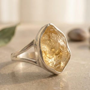 May include: A silver-toned ring featuring a large, raw, translucent yellow gemstone with a faceted, irregular surface. The metal band has a split design that connects to the setting holding the prominent, textured, golden-hued stone.