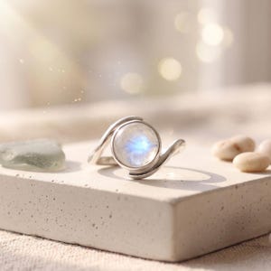 Peut inclure: Une bague en argent ornée d'un cabochon de pierre de lune rond et translucide présentant une adularescence bleue. L'anneau possède une finition lisse et polie avec un design croisé qui entoure la pierre centrale, offrant un aspect épuré.