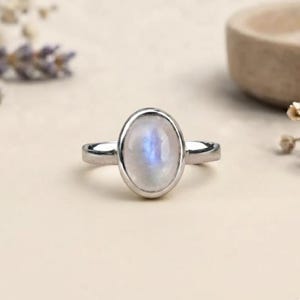 May include: A silver ring featuring a smooth, oval-shaped moonstone cabochon set in a simple metal bezel. The gemstone displays a soft, translucent white appearance with a subtle blue adularescence, centered on a polished silver band.
