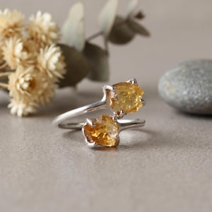 Peut inclure: Une bague ouverte en argent ornée de deux pierres brutes de citrine jaune, maintenues par des griffes métalliques. La bague possède un anneau fin et lisse en argent qui s'enroule autour du doigt, révélant la texture naturelle des pierres.