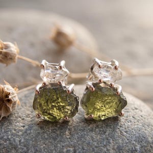 Puede incluir: Un par de pendientes de botón con piedras de moldavita en bruto de color verde oliva situadas bajo cristales de cuarzo transparentes y facetados. Las piedras están sujetas por delicadas garras metálicas, creando un aspecto texturizado.