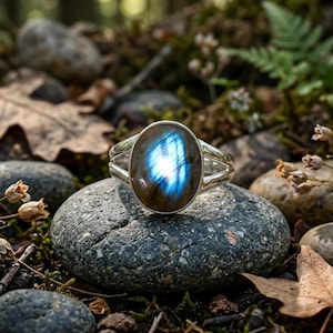 Könnte beinhalten: Ein Silberring mit einem großen, ovalen Labradorit-Edelstein, der einen markanten blauen Schimmer aufweist. Die Ringschiene ist in zwei polierte Metallarme geteilt, die an den Seiten des Steins zusammenlaufen und ein offenes Design bilden.